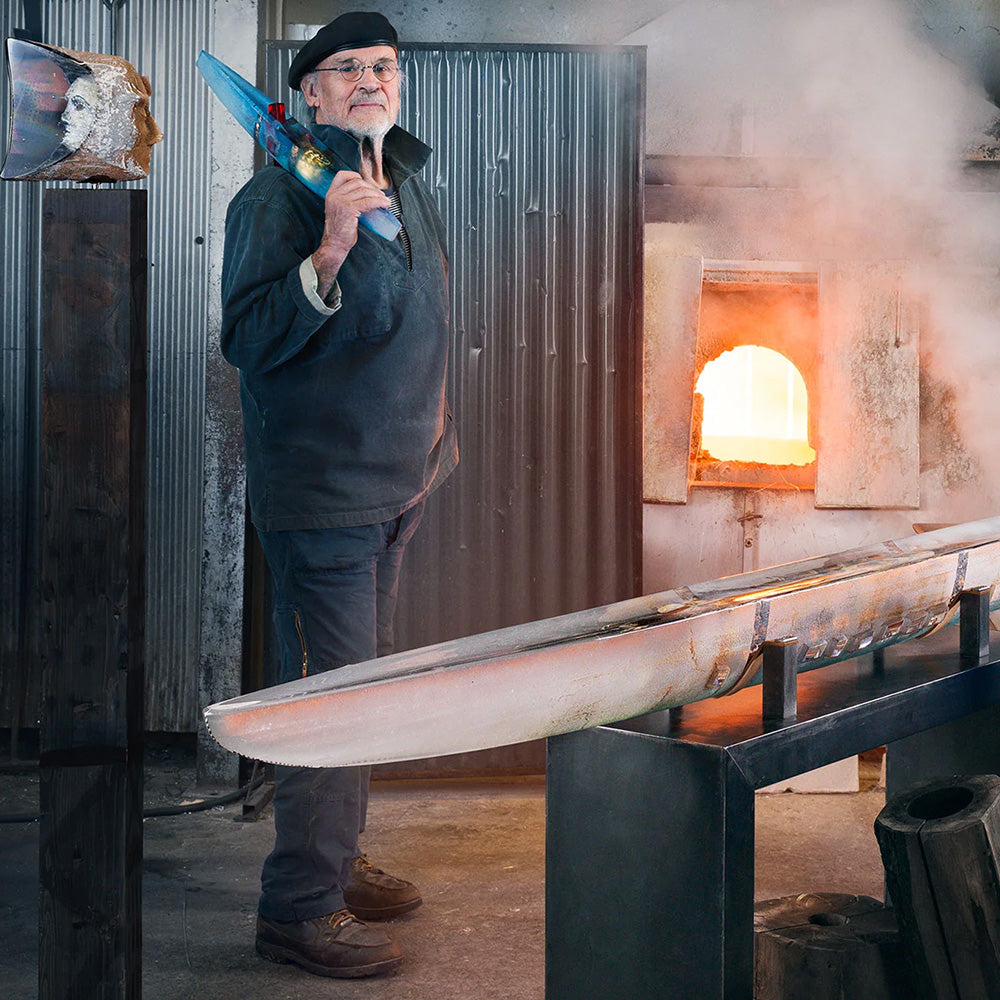 Man working with glass in a furnace, holding a blue art glass boats behind other large art glass sculptures.