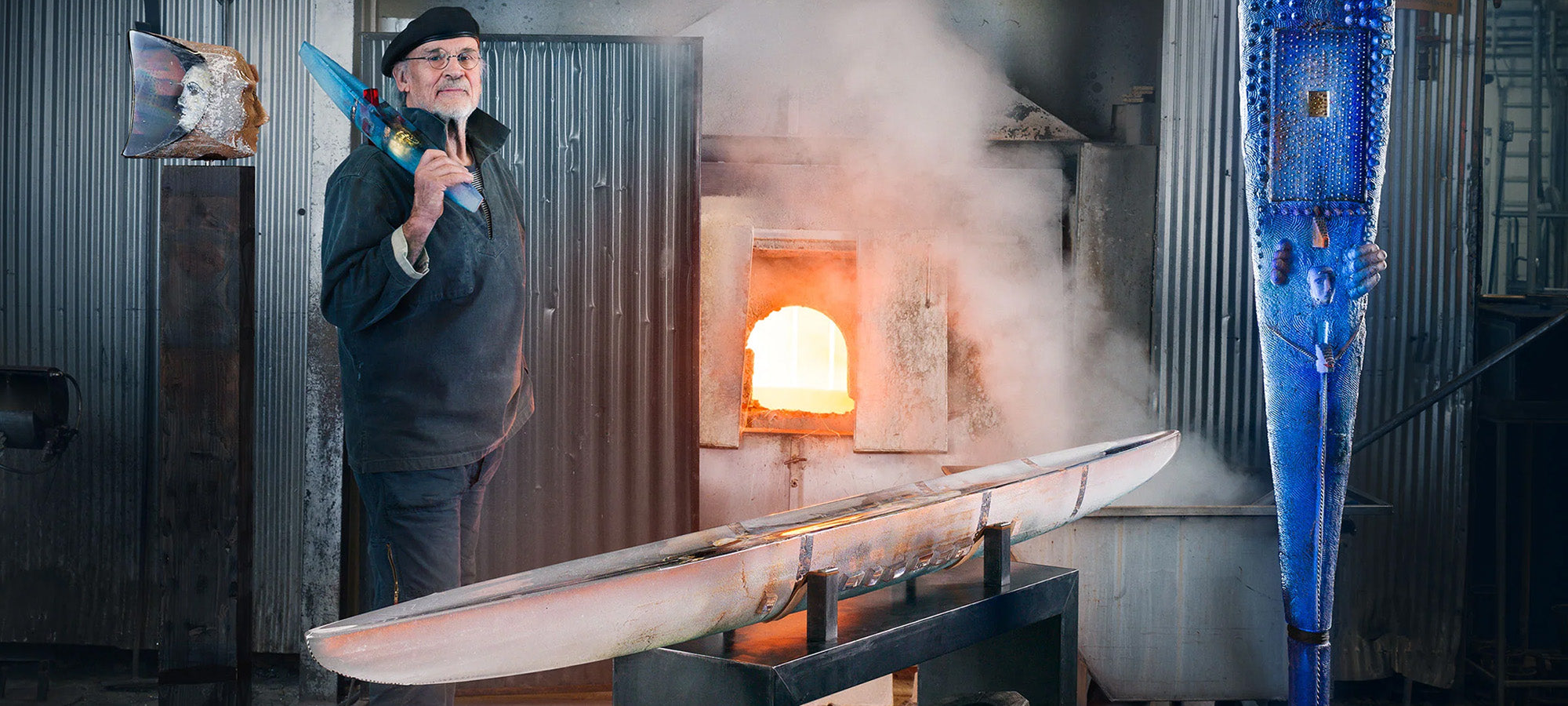 Designer Bertil Vallien holding an art glass boat on his shoulder behind 3 other 3 large art glass sculptures in the Kosta Hot Shop.