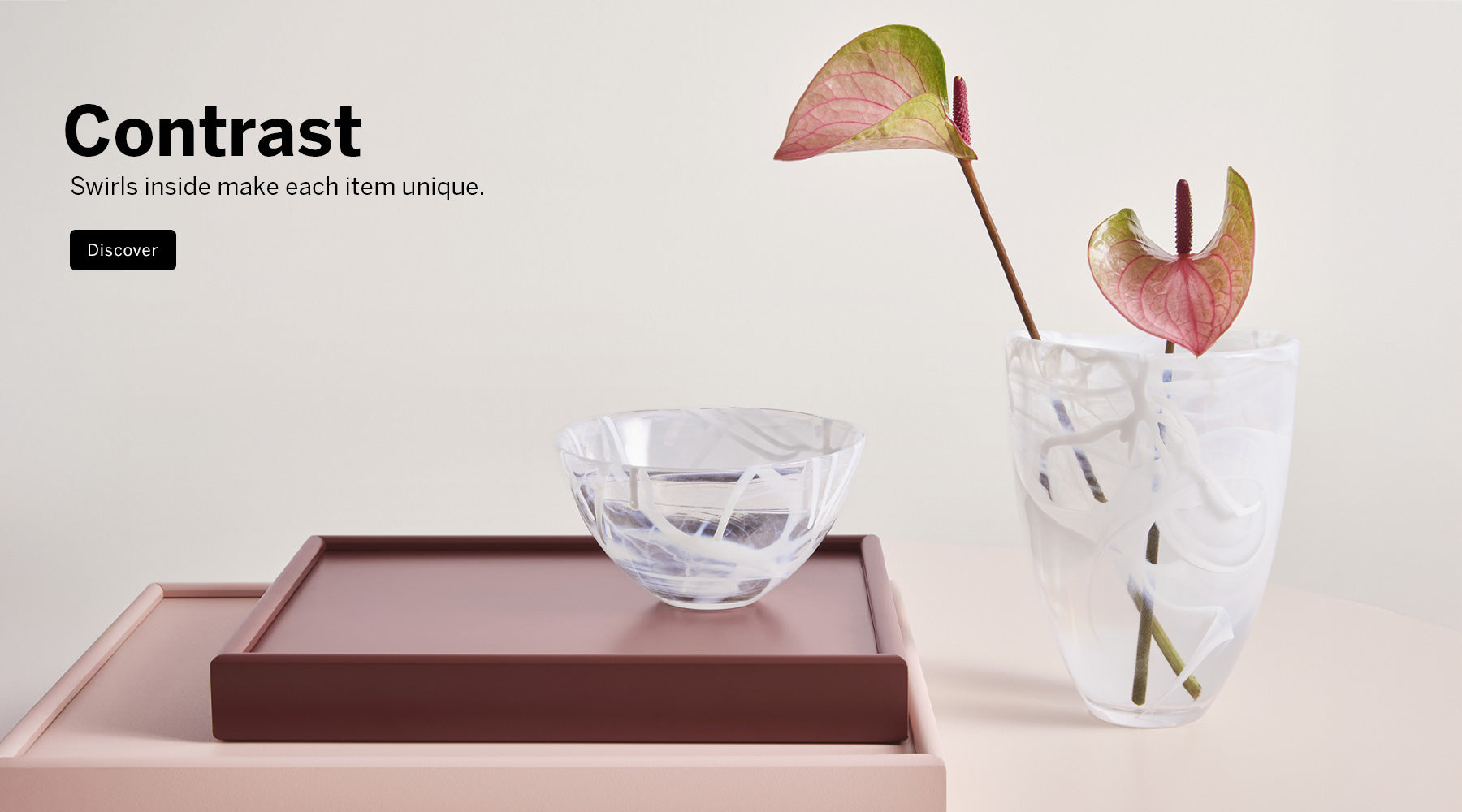 A styled interior scene with crystal Contrast Collection white/White vase and bowl on a dark brown tray and a clear glass vase holding pinkish-red anthurium stems, set against a warm off-white background with the text 'Contrast' Swirls inside make each item unique in the upper left.