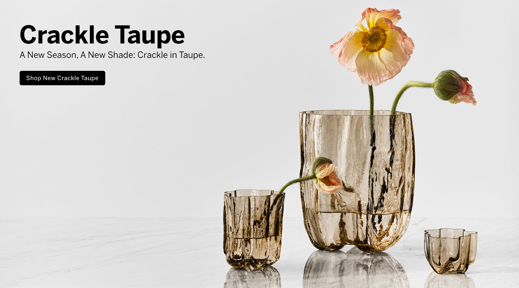 Crackle collection taupe vases and small votive on a marble surface with flower stems in the vases and a light background with text introducing new taupe color for Crackle collection.