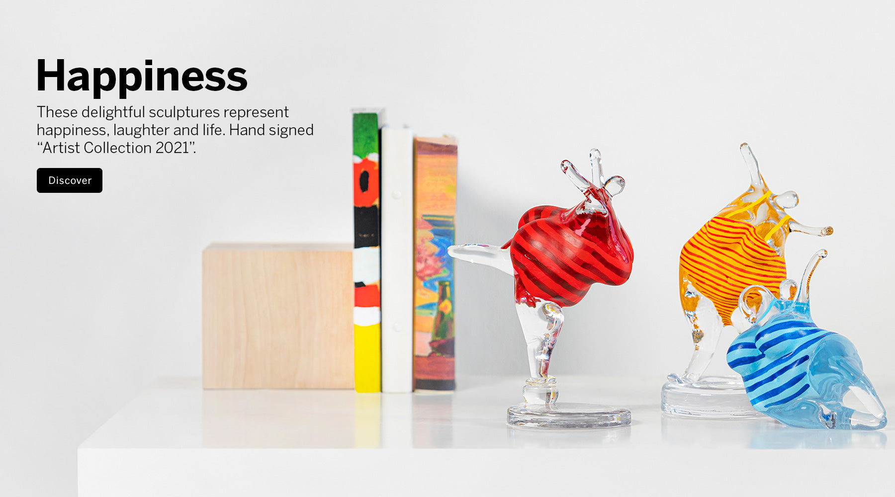 Three Happiness Collection colorful crystal sculptures of robust women in bathing suits on a white surface against a white background, with a wooden block and two books beside them. Text on the left reads 'Happiness' and references the Artist Collection 2021.