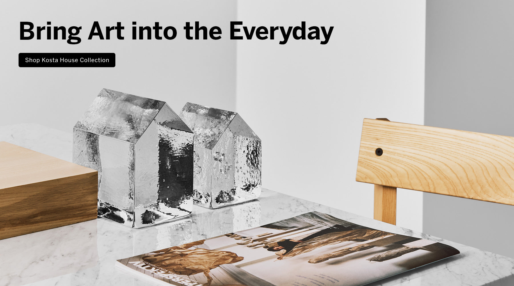 Kosta House collection showing two clear glass house-shaped sculptures with textured faceted sides displayed on a white marble surface next to a light wood chair, with a magazine in front. Text overlay reads 'Bring Art into the Everyday' and 'Shop Kosta House Collection'.