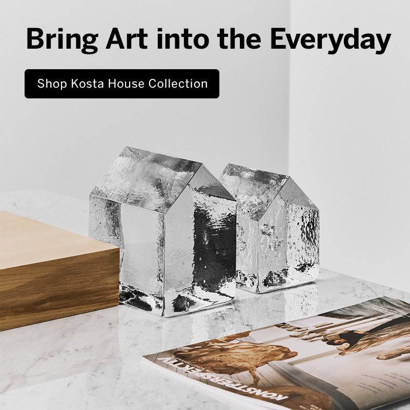 Mobile version image of Kosta House collection showing two clear glass house-shaped sculptures with textured faceted sides displayed on a white marble surface next to a light wood chair, with a magazine in front. Text overlay reads 'Bring Art into the Everyday' and 'Shop Kosta House Collection'.