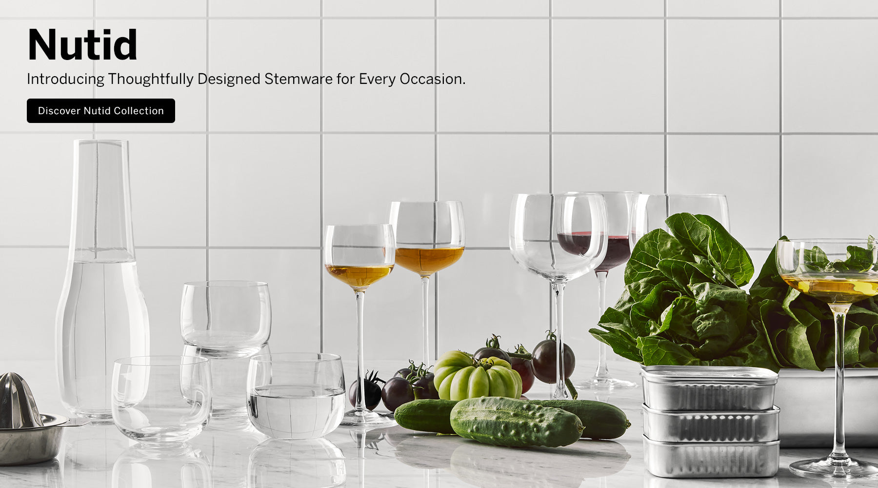 Nutid collection crystal decanter, wine, coupe, apertif, and tumbler glasses on white marble kitchen counter surface with vegetables and a white tile background with text saying 'Introducing Thoughtfully Designed Stemware for Every Occasion, Discover Nutid Collection'.