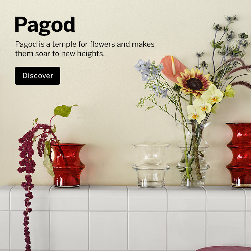 Mobile version image of Pagod Collection red and clear crystal vases in multiple sizes with flowers against in them on tile ledge with a beige wall. Visible text on the left reads 'Pagod' with a short description and a black 'Discover' button.