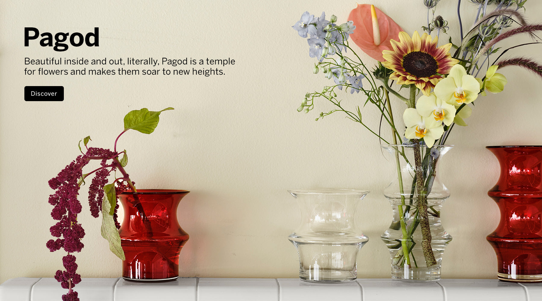 Pagod Collection red and clear crystal vases in multiple sizes with flowers against in them on tile ledge with a beige wall. Visible text on the left reads 'Pagod' with a short description and a black 'Discover' button.