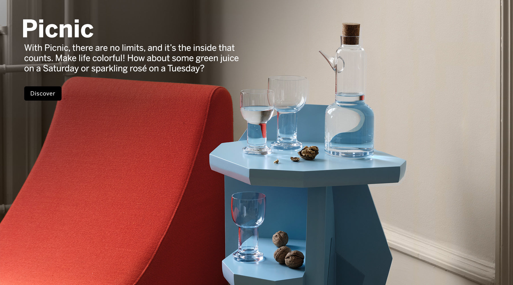 Picnic collection crystal carafe, and glasses on light blue side table with walnuts next to a contemporary orange hair in corner room setting.