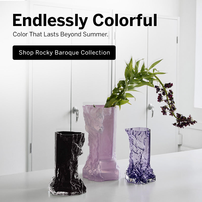 Three textured Rocky Baroque collection glass vases in black, pale lavender, and purple are displayed on a white surface in front of white doors, with a green plant stem inserted into the center container. Large text at the top reads 'Endlessly Colorful' and 'Shop Rocky Baroque Collection.