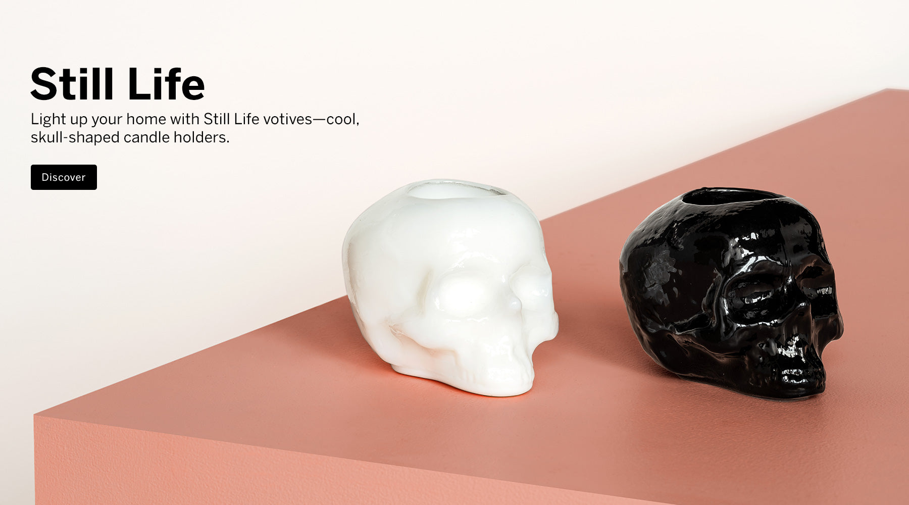 Still Life collection skull shaped white and a black votive candle holder on a peach colored surface and a light background with text promoting Still Life collection.